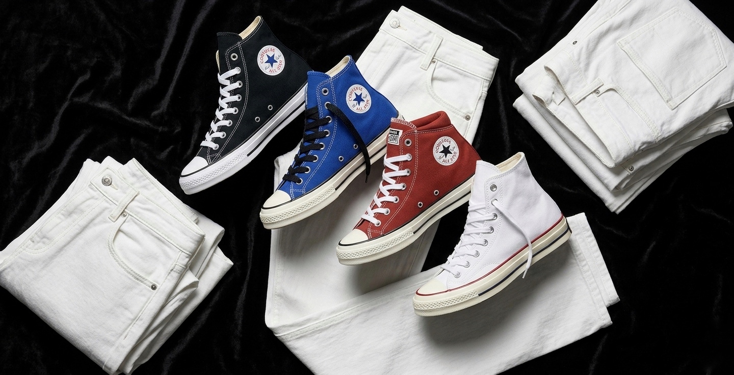 Four pairs of Converse Chuck Taylor All Star sneakers in black, blue, red, and white displayed on folded white denim jeans against a dark fabric background.